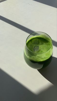 a green smoothie in a glass sitting on the floor next to a shadow cast wall