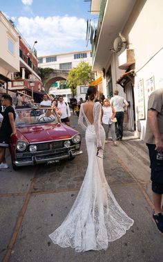 a woman in a wedding dress walking down the street next to an old red car