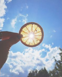 a hand holding an orange in front of a blue sky with white clouds and trees