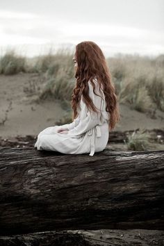 a woman with long red hair sitting on a log in front of some grass and sand