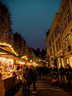 people are walking through an outdoor market with christmas lights on the buildings in the background