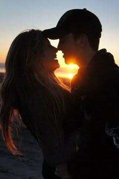 a man and woman are kissing on the beach at sunset with the sun setting behind them