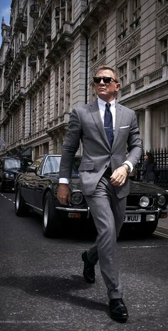 a man in a suit and sunglasses walking down the street next to a black car