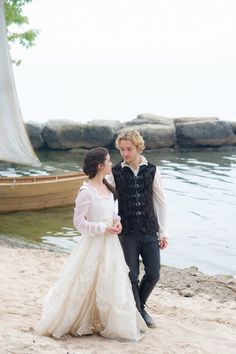 a young man and woman dressed in period clothing walking on the beach next to a boat