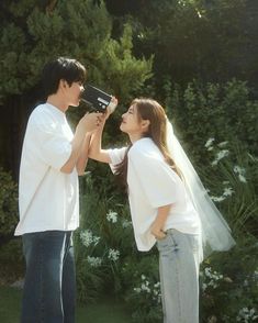 a man and woman standing next to each other in front of bushes holding up a camera