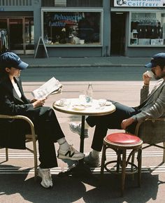 two people sitting at an outdoor table reading newspapers and drinking coffee in front of a store