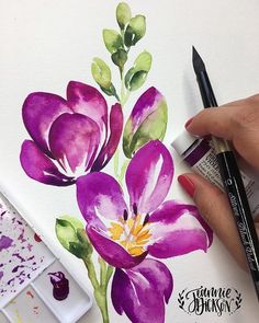 a woman's hand holding a paintbrush over a painting of purple flowers