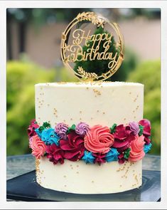 a birthday cake decorated with flowers and a gold happy birthday topper on a table