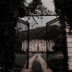 an open gate leading to a large building with a house in the back ground and trees on either side