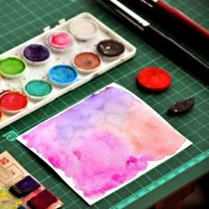 a table with paints, paper and watercolors on it next to a glass of water