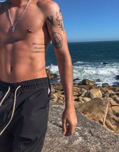 a shirtless man standing on the rocks by the ocean with his hand in his pocket