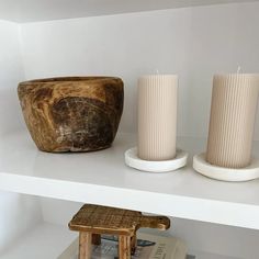 two white candles sitting on top of a shelf next to a wooden bowl and book