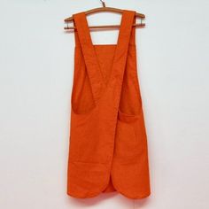 an orange linen vest hanging on a wooden hanger against a white wall with clothes pins attached to it