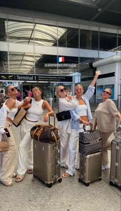 four women are standing with their luggage at the airport and waving to someone in the background