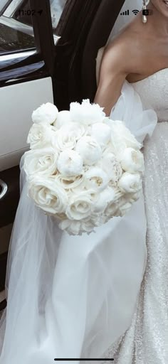 a woman in a wedding dress holding a bridal bouquet next to a white car