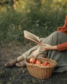 a woman sitting on the ground reading a book next to a basket full of apples