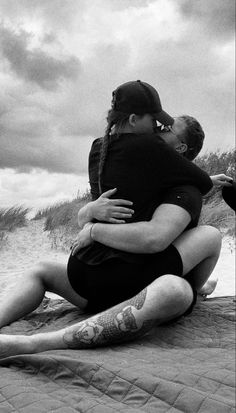 two people sitting on the beach hugging each other and one person has tattoos on his arm