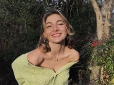 a woman in a green sweater posing for the camera with trees and bushes behind her