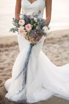 a woman in a wedding dress holding a bridal bouquet on the beach at sunset