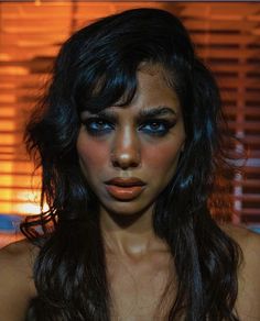 a woman with dark hair and blue eyes posing for a photo in front of blinds