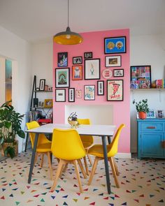 a dining room table with yellow chairs in front of a pink wall and pictures on the wall