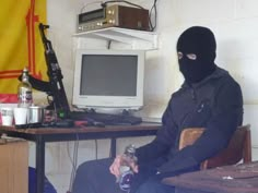 a man in a black mask sitting at a desk with a computer monitor and keyboard