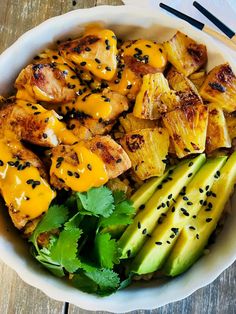 a white bowl filled with chicken, avocado and other vegetables next to chopsticks