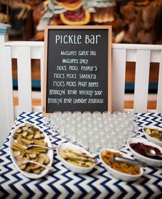 a table topped with lots of food next to a chalkboard sign that says pickle bar