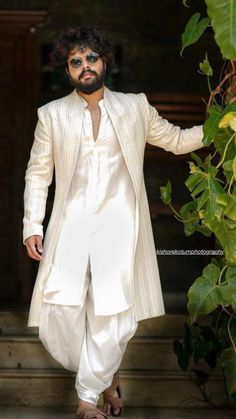 a man in a white suit and sunglasses is standing near some green plants with his hands on his hips