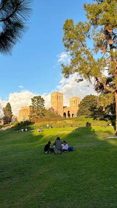 two people are sitting on the grass in front of a large building and some trees