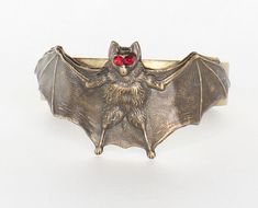 Brass Bat Cuff, made in USA, sold by each This striking brass cuff features a bold design with a bat, its wings dramatically wrapped around the wrist, creating an air of mystery and intrigue. The bat's eyes are adorned with deep red gemstones, adding a captivating and gothic flair. The antique brass finish enhances its vintage appeal, while the intricate detailing of the wings and bat make it a unique statement piece, perfect for those with a love for dark, edgy accessories. Dimensions: 37.8mm x Bat Eyes, Edgy Accessories, Brass Cuff, Brass Bracelet, Red Gemstones, The Wings, Brass Finish, Deep Red, Antique Brass