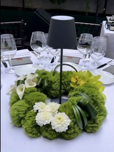 a green wreath with white flowers and greenery sits on a table in front of wine glasses