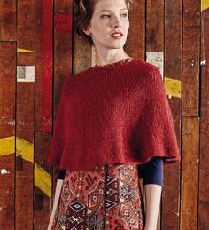 a woman standing in front of a wooden wall wearing a red sweater and patterned skirt