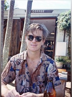 a man sitting at a table with a beer in front of him and palm trees behind him