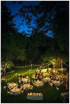 a group of people sitting at tables in the middle of a field with lights strung around them