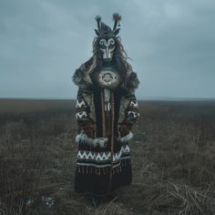 a person in an animal mask and coat standing in a field with tall grass on a cloudy day