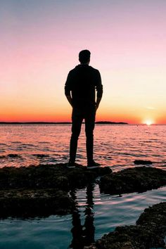 a man standing on top of a rock near the ocean at sunset with his back to the camera