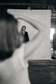 a woman standing in front of a white wall with her hands behind her head looking into the mirror