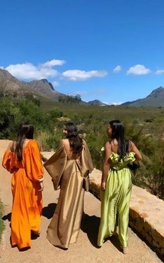 three women are walking down a path in long dresses, with mountains in the background