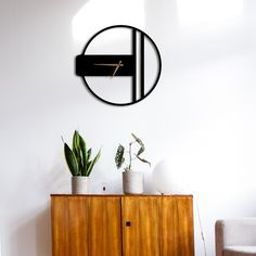 a clock mounted to the side of a white wall next to a wooden cabinet with potted plants on top