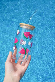 a hand holding a watermelon tumbler next to a swimming pool with flowers on it