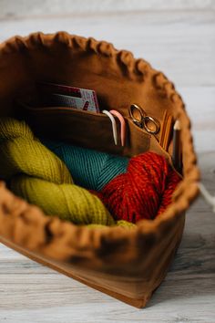 a bag with yarn and knitting needles in it on a wooden table next to a pair of glasses