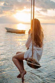 a woman is sitting on a swing in the water with boats behind her at sunset