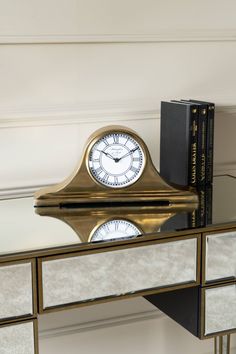 a gold clock sitting on top of a glass table next to a book shelf and mirror