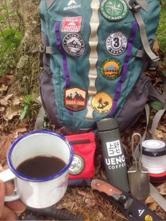 a backpack with patches on it sitting in the woods next to a cup of coffee