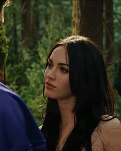 a woman standing next to a man in a forest looking at each other with trees behind her