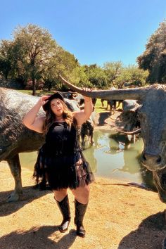 A woman dressed in a black dress, boots, and hat stands beside a bronze longhorn statue, holding its horn. The scene is set in a sunny outdoor area with more statues, a reflective pond, and trees in the background.