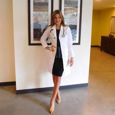 a woman standing in front of a wall wearing a white coat and black dress with gold heels