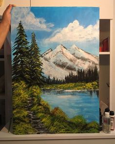 a person holding up a painting in front of a white wall with trees and mountains on it