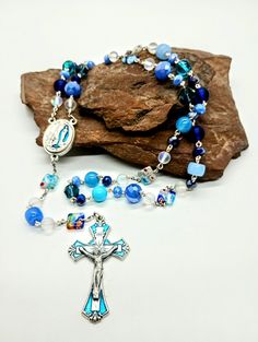 Ladies and Gentlemen, may I introduce you to the extraordinary Obstinate Blue: Ooo do we have a good one for you here today people!  Another named piece that just shines and shimmers with personality. A captivating 21-inch-long rosary meticulously crafted from blue lampwork and glass beads in a vibrant hodgepodge style. This spirited and unyielding prayer tool boldly asserts itself into your life draped in brilliant shades of azure. As an advocate for your desires and needs this rosary is second to none, it invites you to embrace a unique and defiant approach to your spirituality that will not only benefit you but the people around you. Now, the name Obstinate Blue may raise a few eyebrows when associated with a rosary. The notion of obstinacy, often seen as stubbornly refusing to change o Handmade Blue Crucifix Jewelry, Rosary Art, Blue Rosary, Power Of Meditation, Meditation Beads, Professional Growth, Ladies And Gentlemen, Spiritual Connection, Lady And Gentlemen